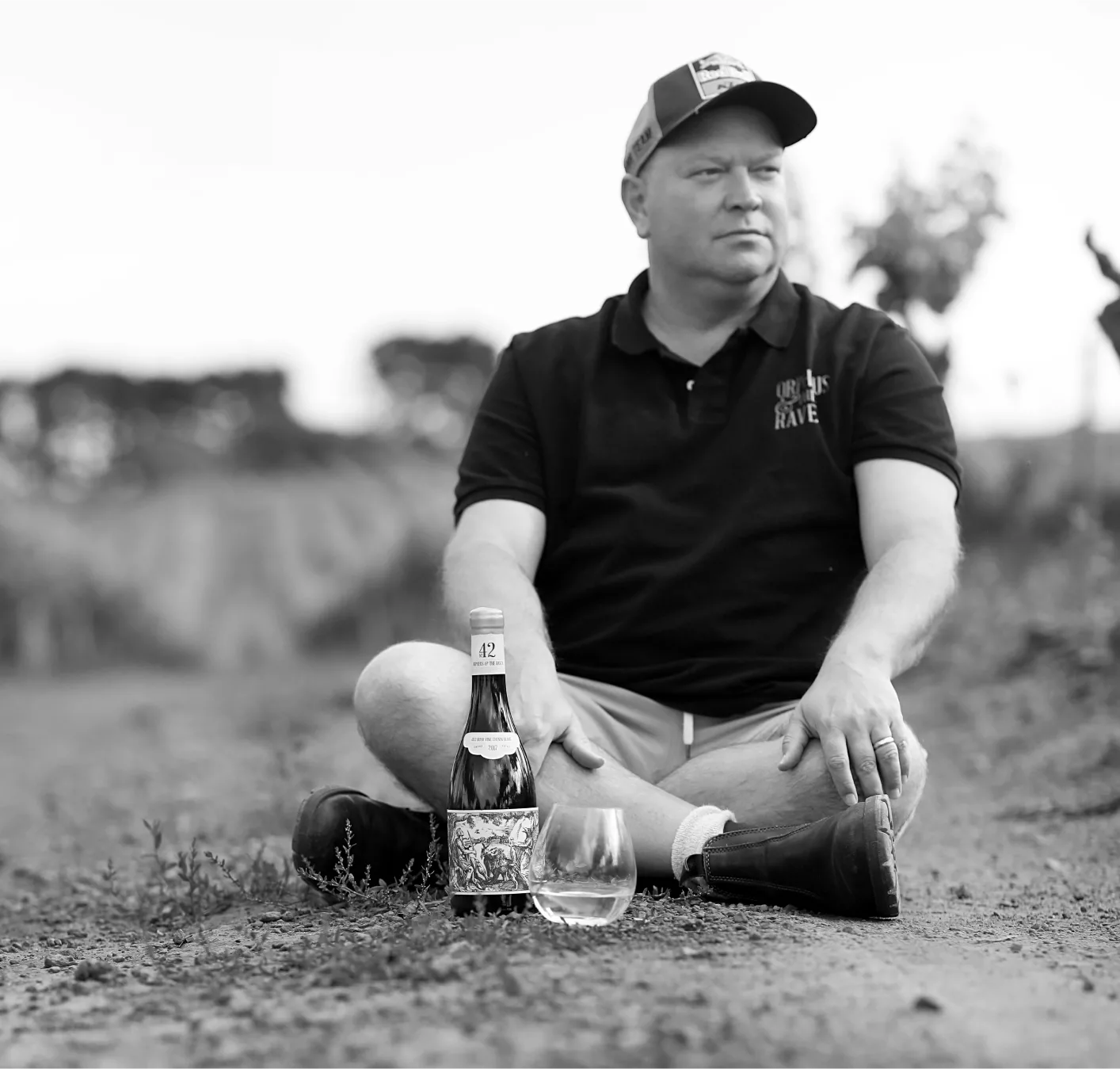 Black and white image of man sitting cross-legged in vineyard with bottle of No. 42 Cape Blend wine