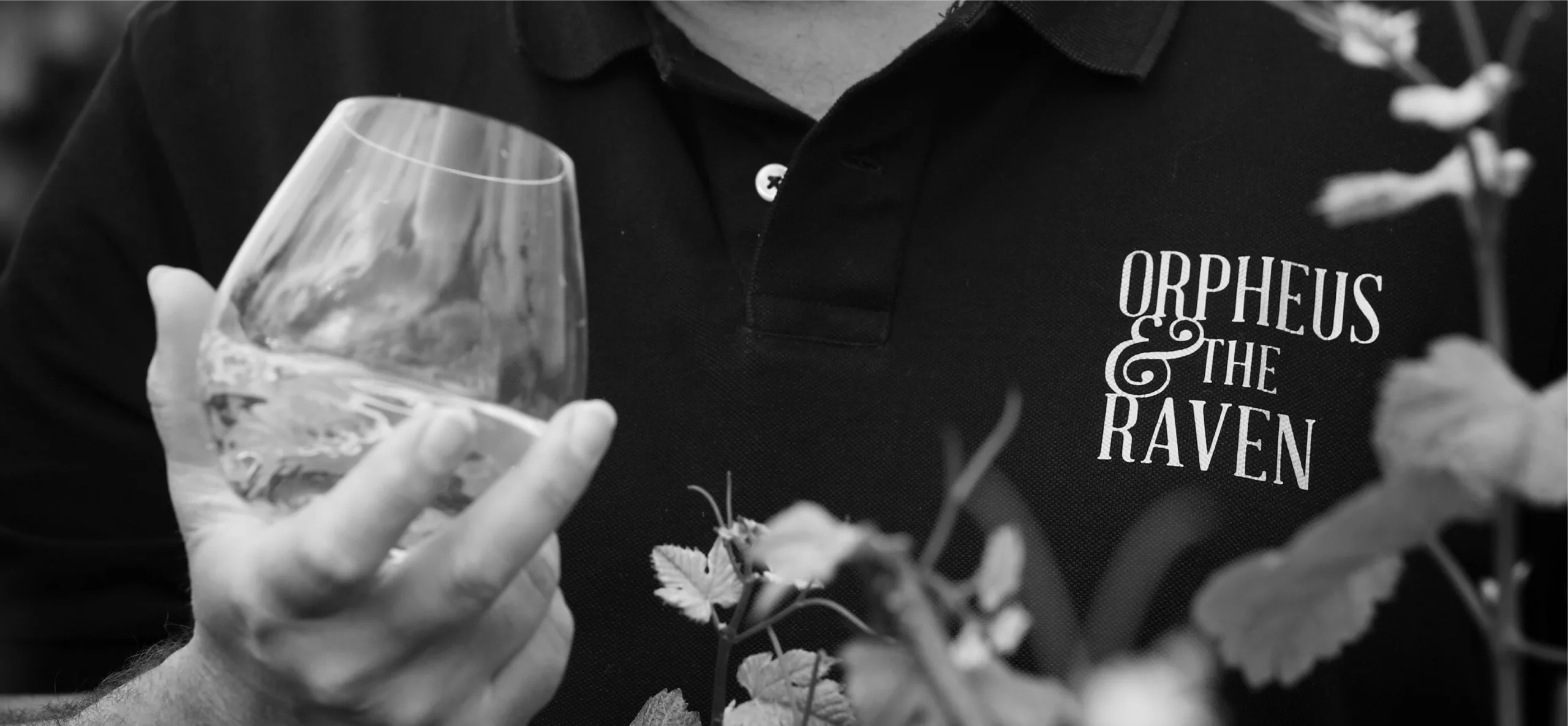Man wearing black button-up golf shirt with Orpheus & The Raven logo, holding wine tumbler
